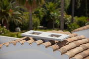 Base doble de hormigon color blanco con dos aberturas rectangulares instalada sobre el tejado de teja curva de una villa mediterranea, jardin con palmeras desenfocado al fondo, luz de mediodia