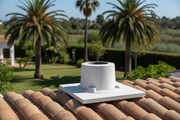 Base cuadrada de hormigon color blanco con abertura circular central instalada sobre el tejado de teja curva de una villa mediterranea blanca, alrededor de la salida de humos, jardin con palmeras desenfocado al fondo, luz de mediodia